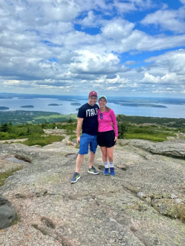 Two people stand on a rocky hilltop with greenery and water islands visible in the background under a blue sky with scattered clouds.