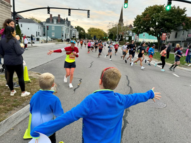 Runners race down a city street past cheering spectators, including two children in blue jackets standing near the curb with arms outstretched.