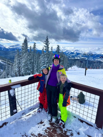 Three people in ski gear stand on a snowy deck with mountains, trees, and ski lifts in the background under a partly cloudy sky.