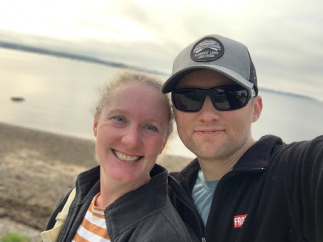 Two people wearing jackets are smiling at the camera while standing on a rocky beach with water and a cloudy sky in the background.