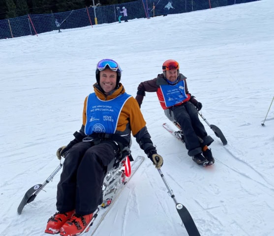 Two people using adaptive ski equipment and wearing blue vests are sitting on a snowy ski slope, holding outriggers, with a ski fence and trees in the background.