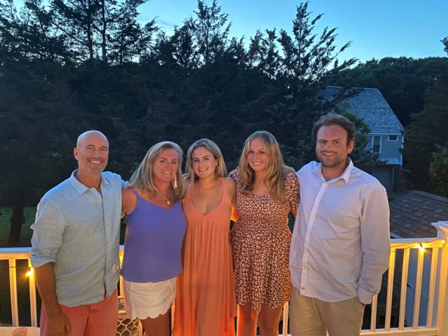 Five people stand together and smile on a porch at sunset, with trees and a house in the background.