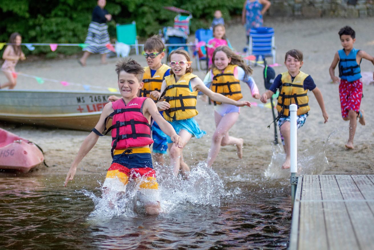 A group of children run into the water with swimming vests on.