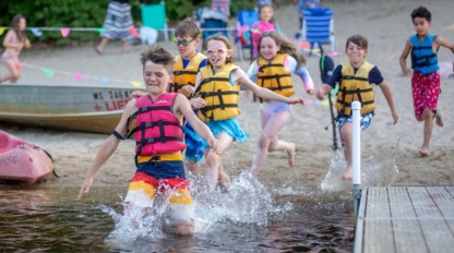 A group of children run into the water with swimming vests on.