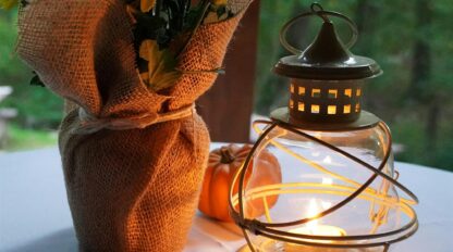 A glass lantern with a lit candle sits on a table next to a burlap-wrapped bouquet of yellow flowers and a small pumpkin, with greenery visible in the background.