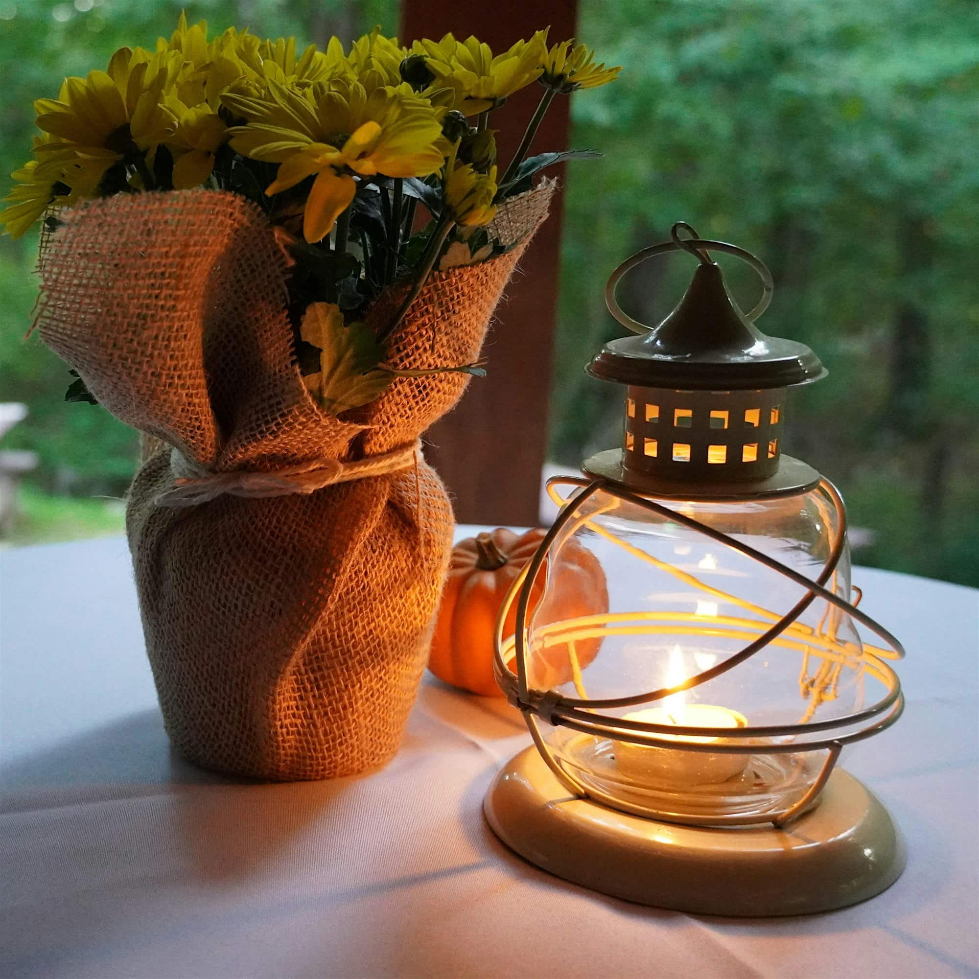 A glass lantern with a lit candle sits on a table next to a burlap-wrapped bouquet of yellow flowers and a small pumpkin, with greenery visible in the background.