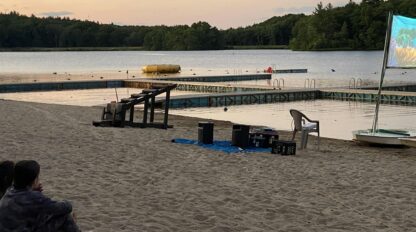 Two people sit on a sandy beach at dusk, watching a movie projected on a screen near a dock with a lake and trees in the background.
