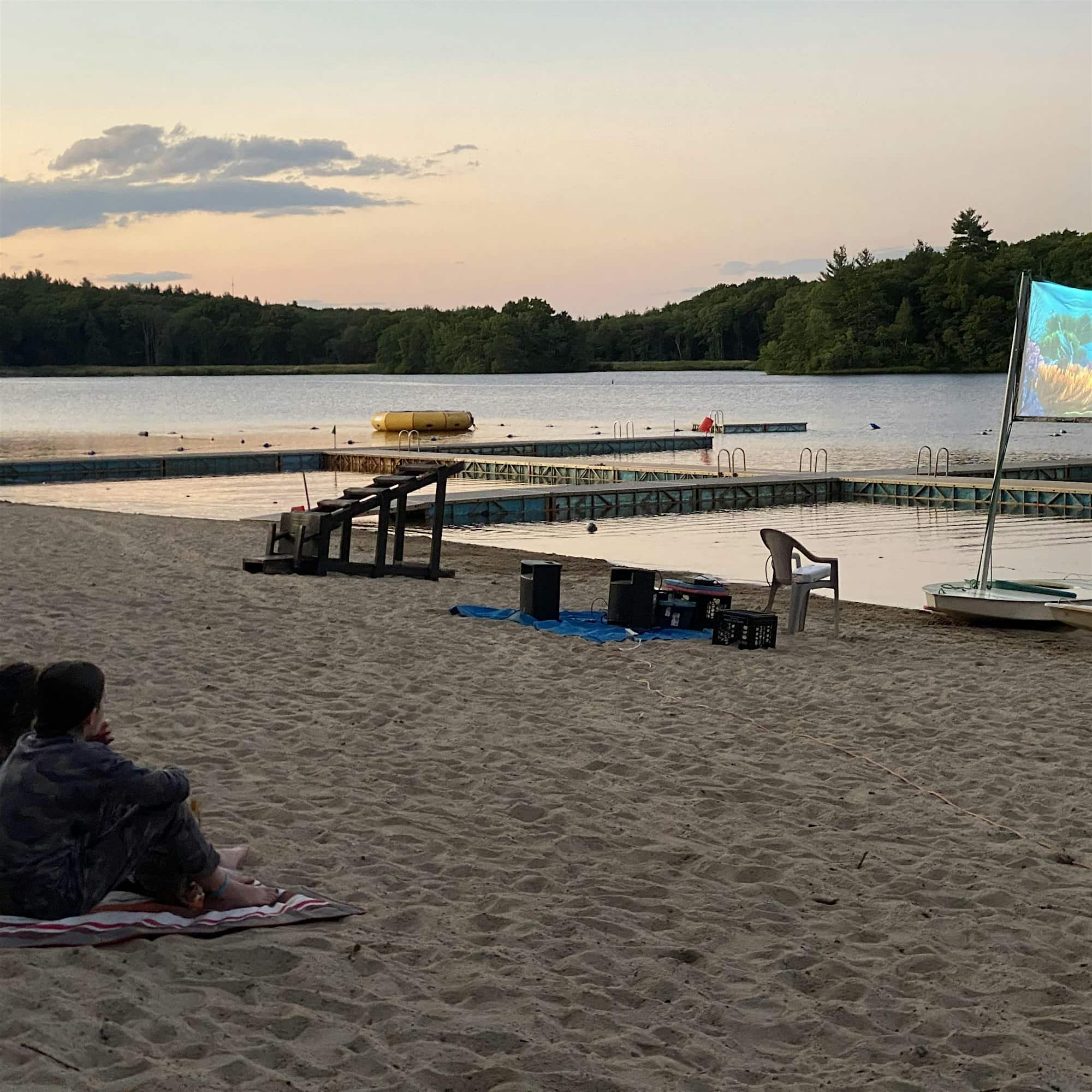 Two people sit on a sandy beach at dusk, watching a movie projected on a screen near a dock with a lake and trees in the background.