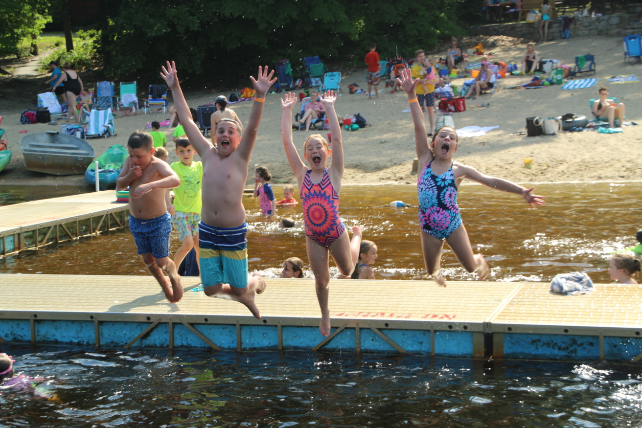 Four children in swimwear jump off a dock into a lake with arms raised, while people relax on a sandy beach with trees and chairs in the background.