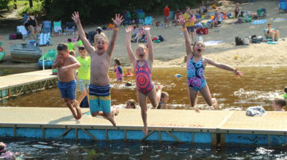 Four children in swimwear jump off a dock into a lake with arms raised, while people relax on a sandy beach with trees and chairs in the background.