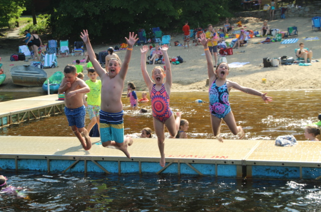 Four children in swimwear jump off a dock into a lake with arms raised, while people relax on a sandy beach with trees and chairs in the background.