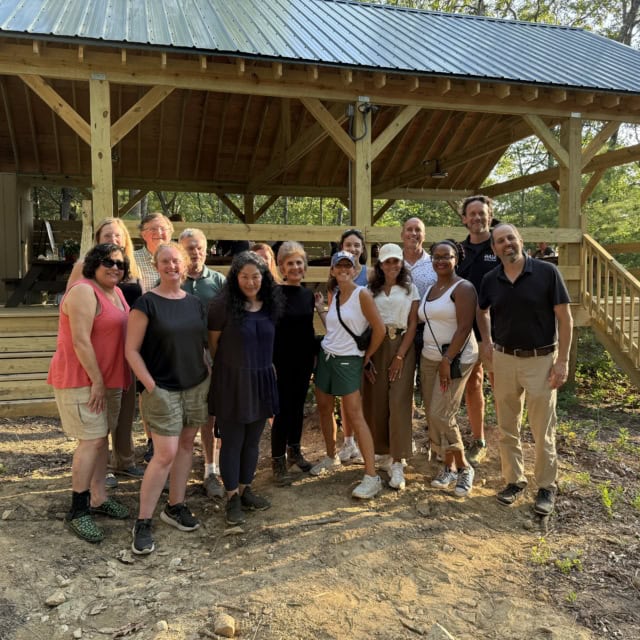 A group of people pose for a photo outdoors in front of a wooden pavilion, with trees and sunlight in the background.