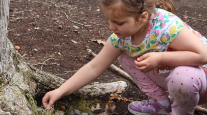 A young girl in patterned clothes crouches by a tree, reaching toward the ground, in a wooded outdoor area.