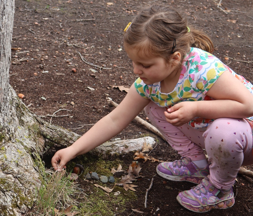 A young girl in patterned clothes crouches by a tree, reaching toward the ground, in a wooded outdoor area.