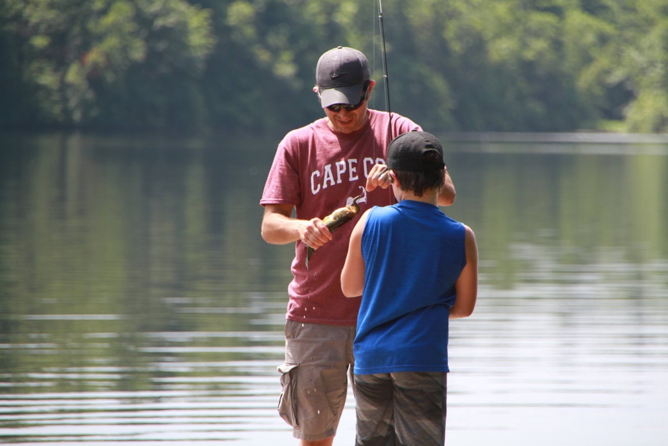 An adult and a child stand by a calm pond. The adult, holding a fishing rod, shows the child a small fish they have caught. Both wear caps; trees are visible in the background.