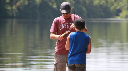 An adult and a child stand by a calm pond. The adult, holding a fishing rod, shows the child a small fish they have caught. Both wear caps; trees are visible in the background.