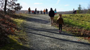 A group of people, including adults and children, walk up a gravel trail on a sunny day with clear blue skies and scattered clouds.