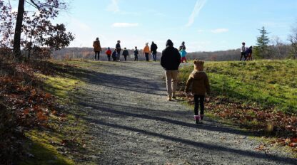 A group of people, including adults and children, walk up a gravel trail on a sunny day with clear blue skies and scattered clouds.