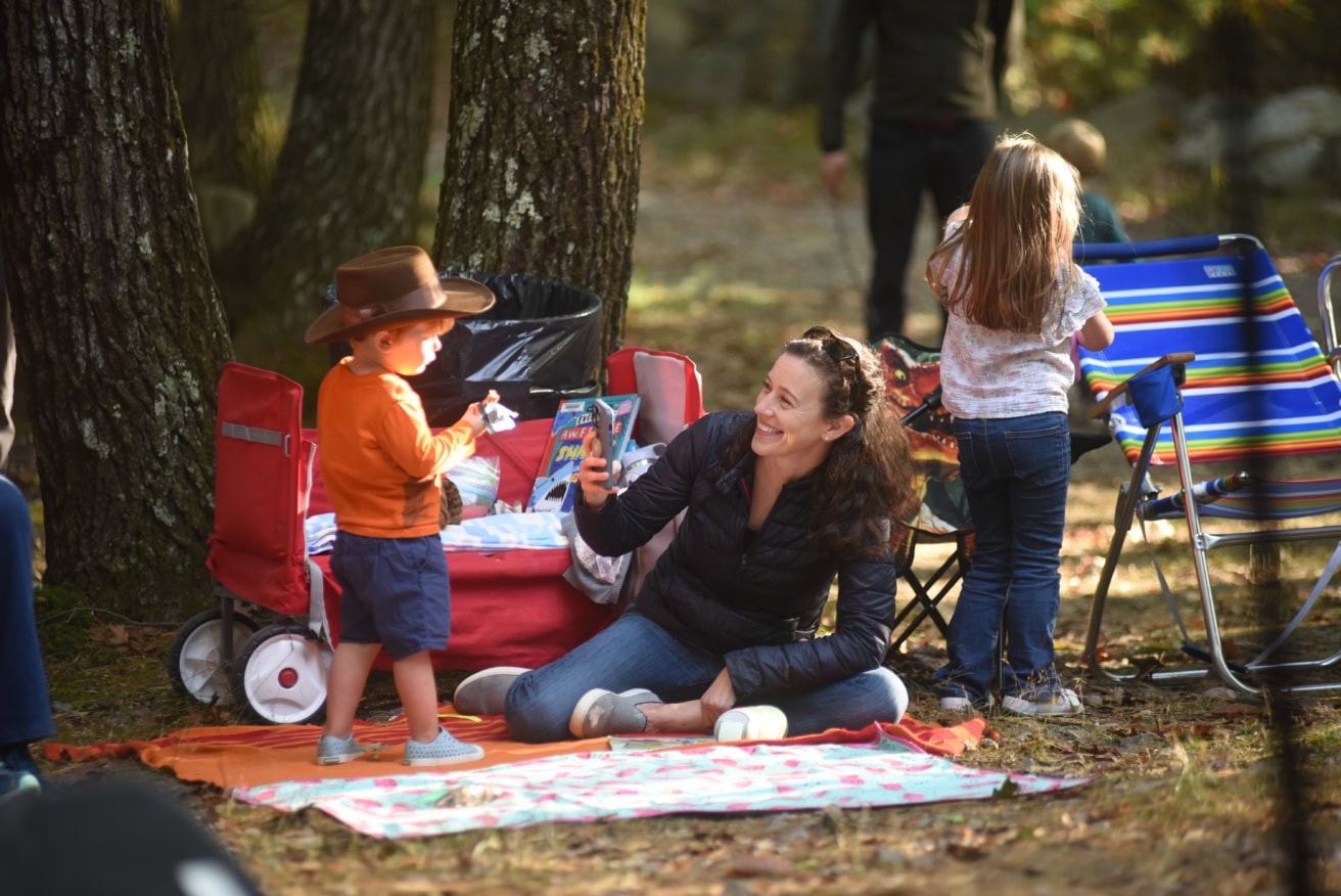 A woman sits on a picnic blanket taking a photo of a young boy wearing a brown hat, while a girl stands nearby next to a colorful folding chair in a wooded outdoor setting.