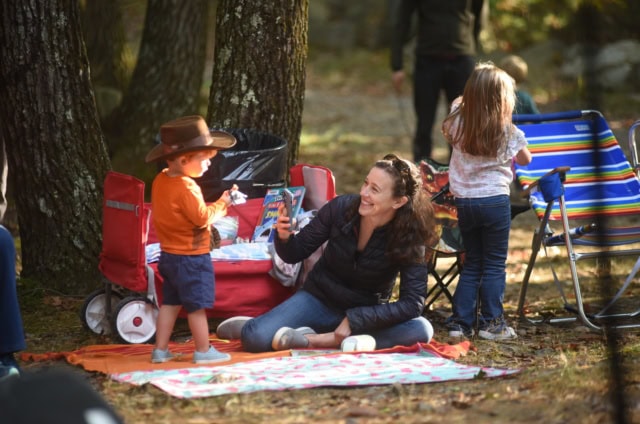 A woman sits on a picnic blanket taking a photo of a young boy wearing a brown hat, while a girl stands nearby next to a colorful folding chair in a wooded outdoor setting.