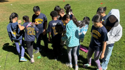 A group of children stands closely together on a grassy field, many wearing blue shirts labeled "Farmington Kickball." Some face away while others look down.