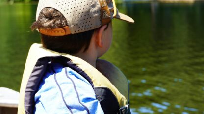 A child wearing a life jacket and cap sits in a boat, looking out over a calm lake with trees in the background.