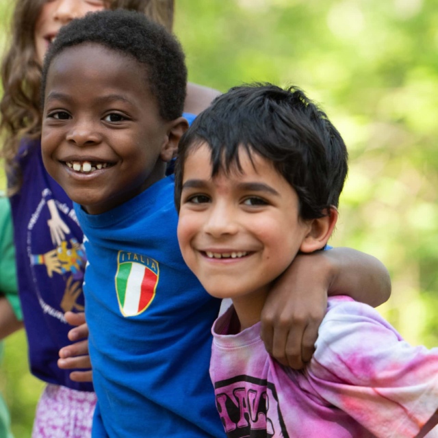Three smiling children stand together outdoors with their arms around each other's shoulders.