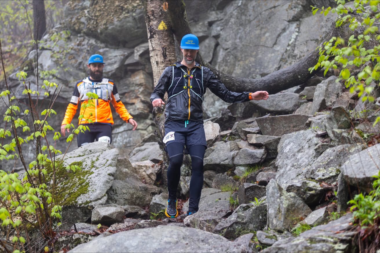 Two people in athletic gear and blue hats carefully descend a rocky, uneven trail surrounded by trees and large rocks during a trail race.