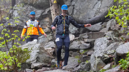 Two people in athletic gear and blue hats carefully descend a rocky, uneven trail surrounded by trees and large rocks during a trail race.