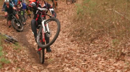 A group of cyclists ride on a leaf-covered forest trail, with one person in front performing a wheelie.