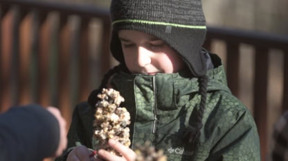 A child wearing a winter coat and hat holds a pinecone covered in seeds, standing outdoors near a wooden railing.