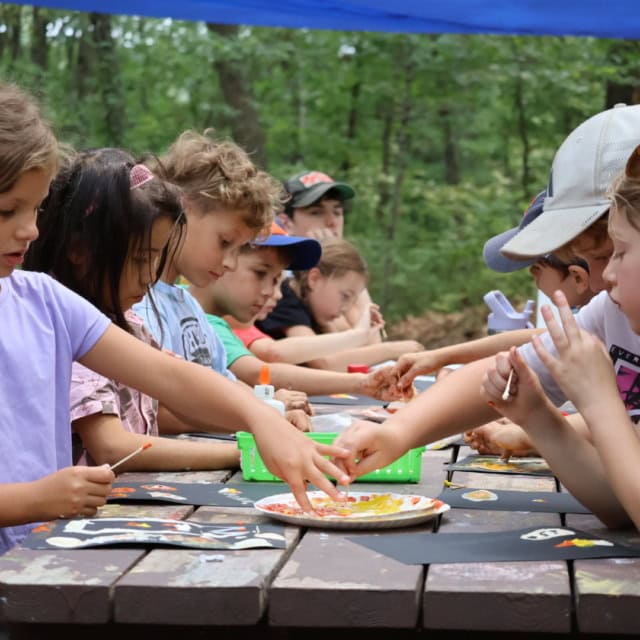A group of children sit at a picnic table outdoors, engaged in arts and crafts activities with paint, brushes, and paper.