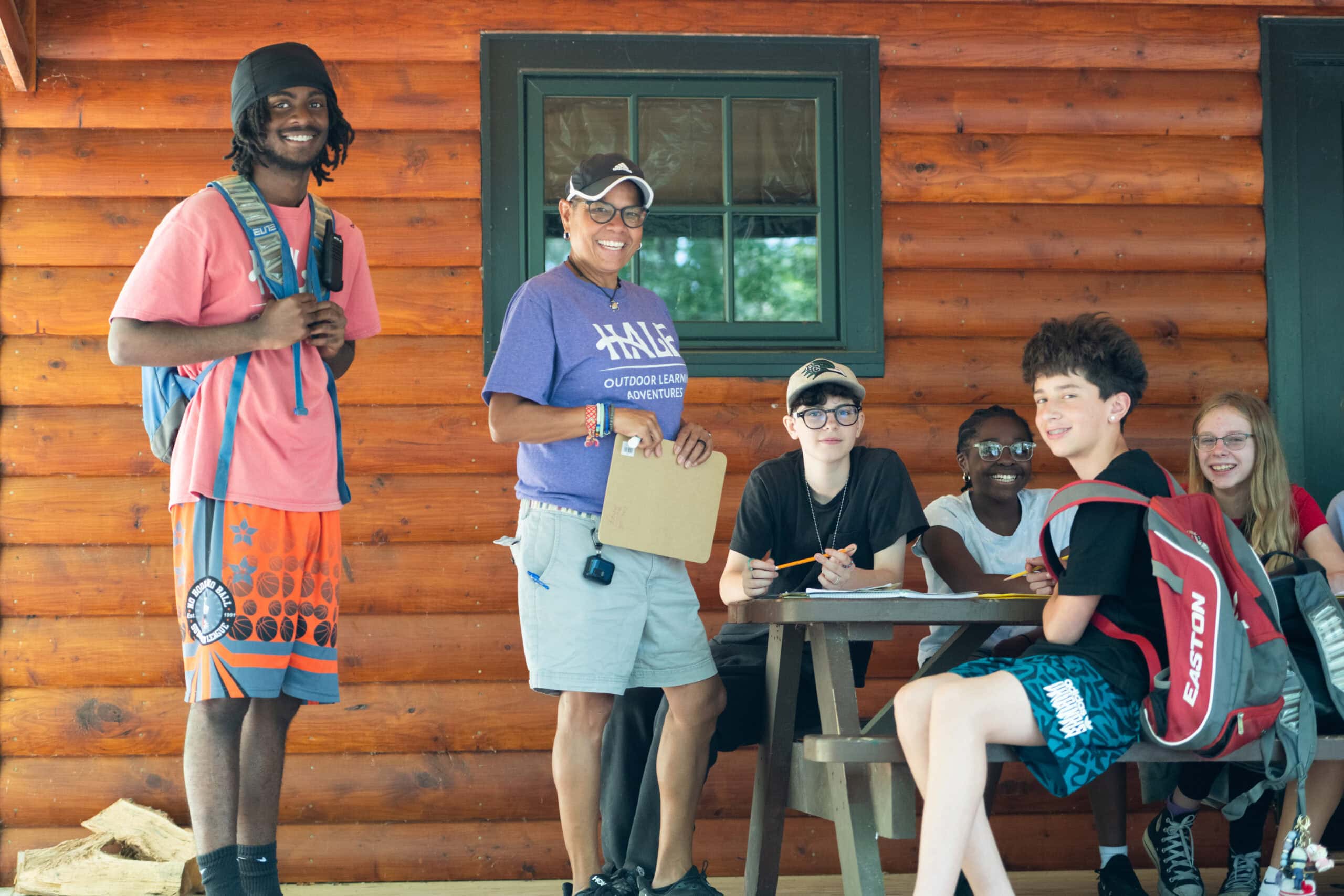 A group of young people and an adult stand and sit together outside a wooden building, some holding pencils and notebooks, smiling at the camera.