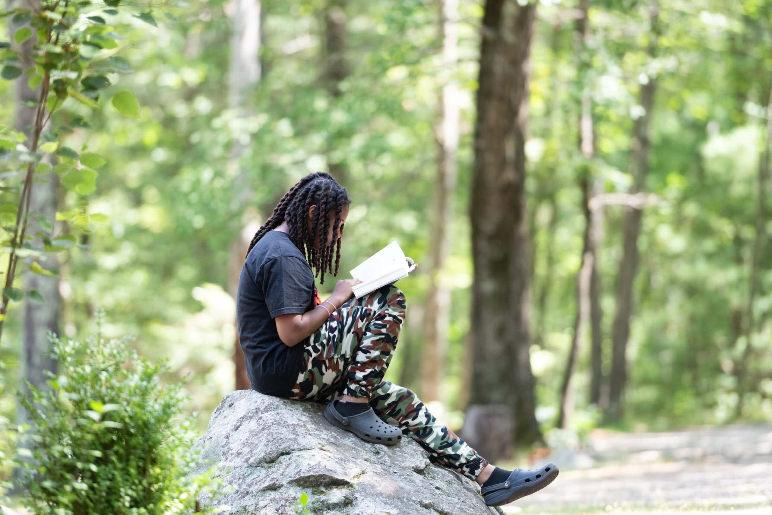 Person with long braids sits on a large rock in the woods, wearing a black shirt, camouflage pants, and Crocs, reading a book.