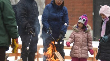 Four people and two children toast marshmallows over an outdoor fire pit in winter, with snow on the ground and a brick building in the background.