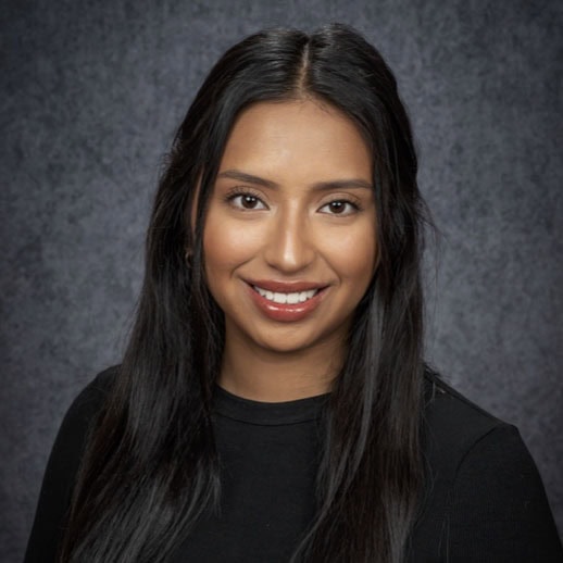 A woman with long dark hair, wearing a black top, smiles at the camera against a mottled gray background.