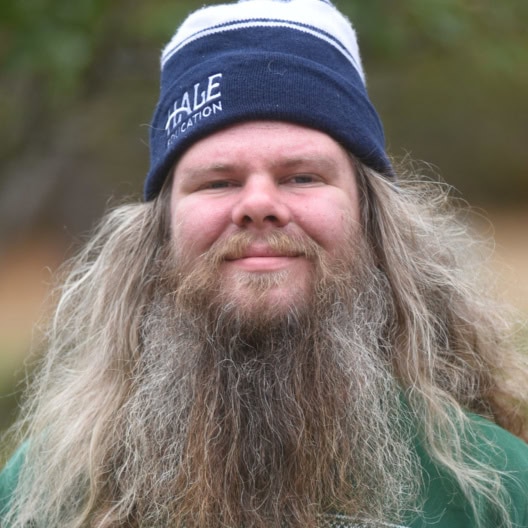 A man with long gray hair and a full beard wears a blue and white knit hat and a green sweater, standing outdoors.