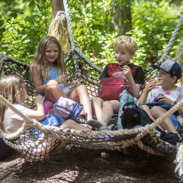 Four children sit on a large rope hammock in a wooded area, talking and eating snacks with backpacks around them.