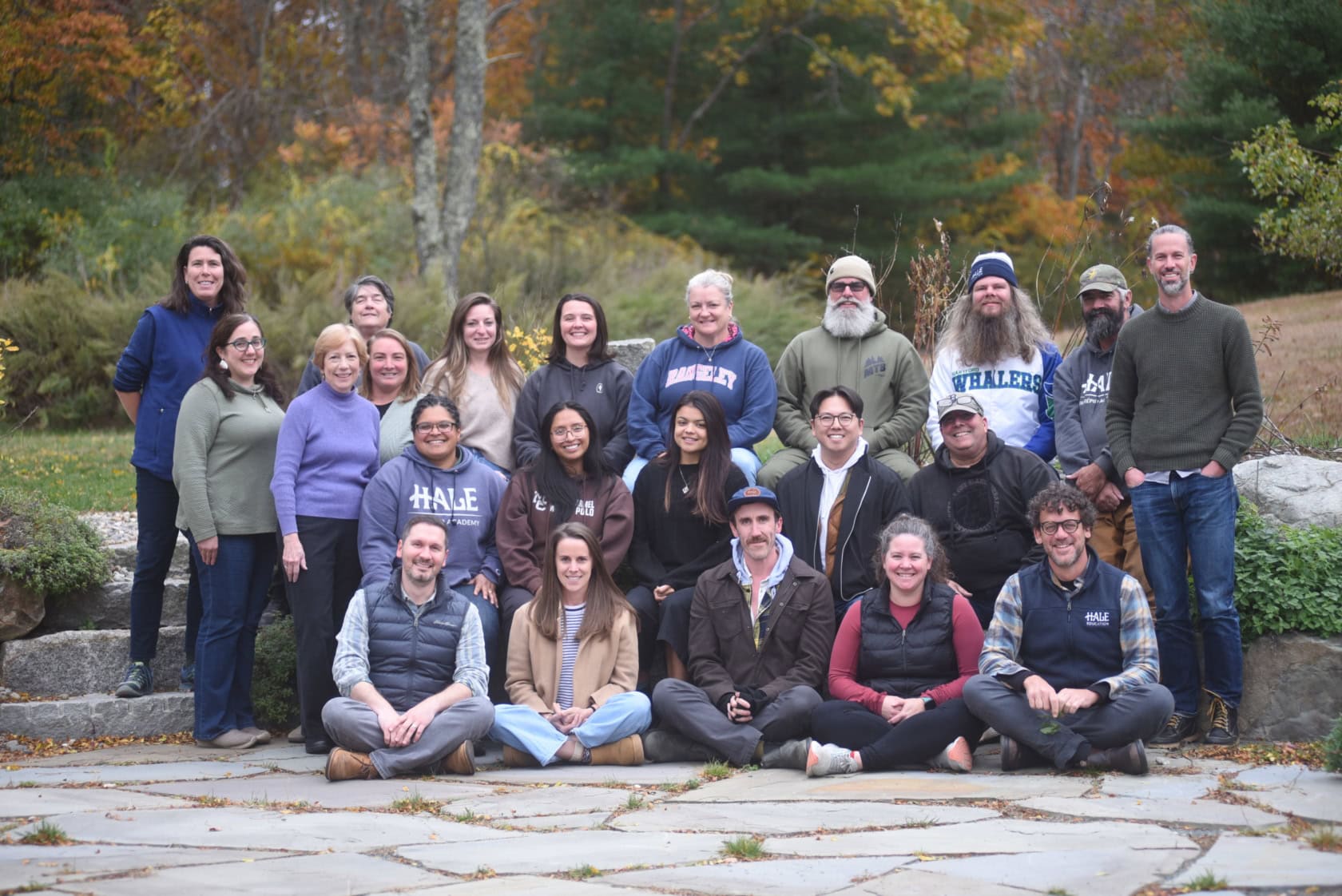 A group of 21 adults poses outdoors on a stone patio, with autumn trees and greenery in the background. Some are seated while others stand behind them, all facing the camera.