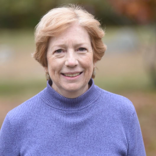 A woman with short, light brown hair wears a purple sweater and smiles at the camera outdoors, with a blurred natural background.
