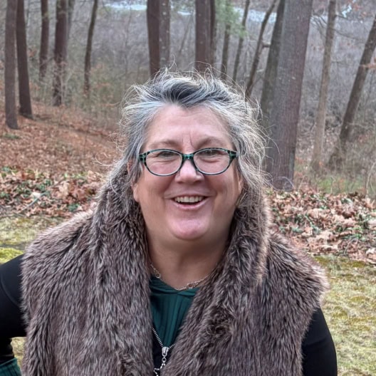 A woman with gray hair and glasses, wearing a faux fur vest, stands outdoors in a wooded area, smiling at the camera.