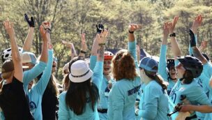 A group of people in blue shirts and helmets raise their arms in celebration outdoors, surrounded by trees on a sunny day.