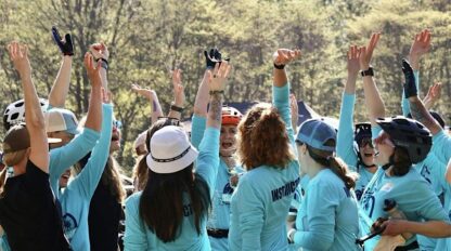 A group of people in blue shirts and helmets raise their arms in celebration outdoors, surrounded by trees on a sunny day.