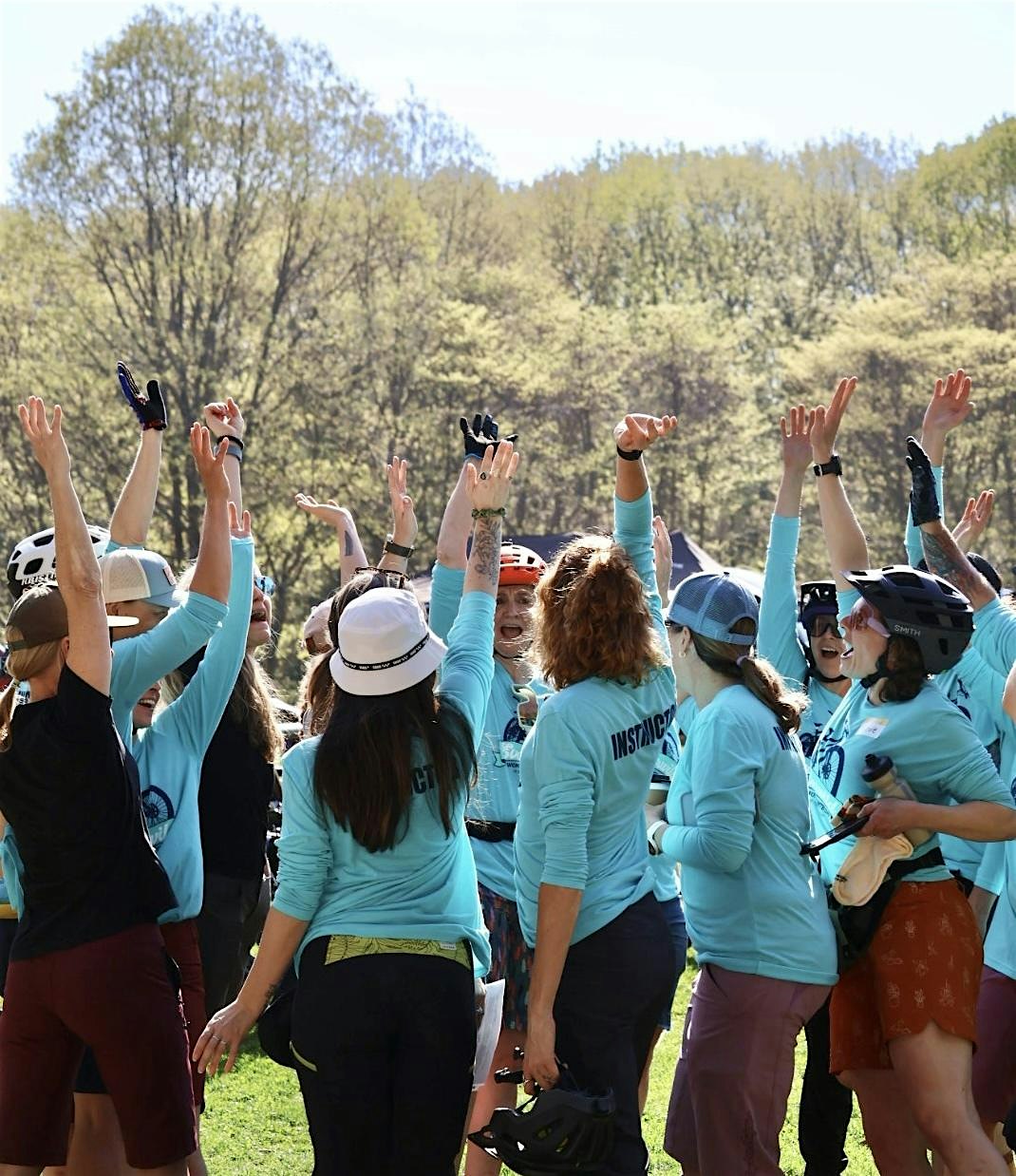 A group of people in blue shirts and helmets raise their arms in celebration outdoors, surrounded by trees on a sunny day.