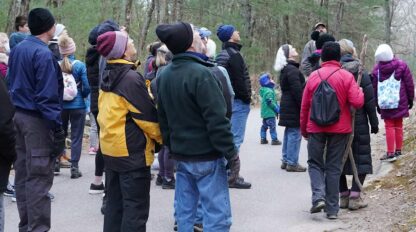 A group of people in outdoor clothing stand on a paved path in a forested area, looking in the same direction, possibly listening to a guide.
