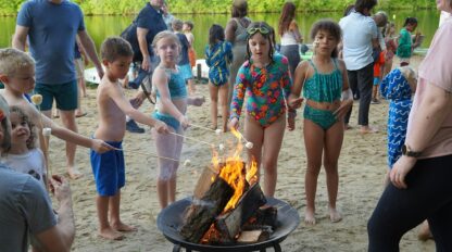 Children in swimsuits toast marshmallows over a fire pit on a sandy beach, with adults and other kids in the background near trees and water.