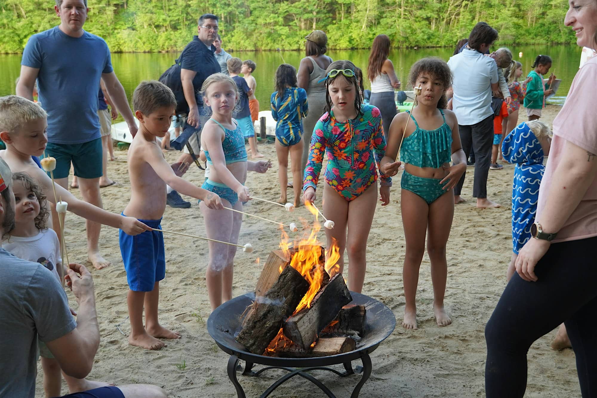 Children in swimsuits toast marshmallows over a fire pit on a sandy beach, with adults and other kids in the background near trees and water.