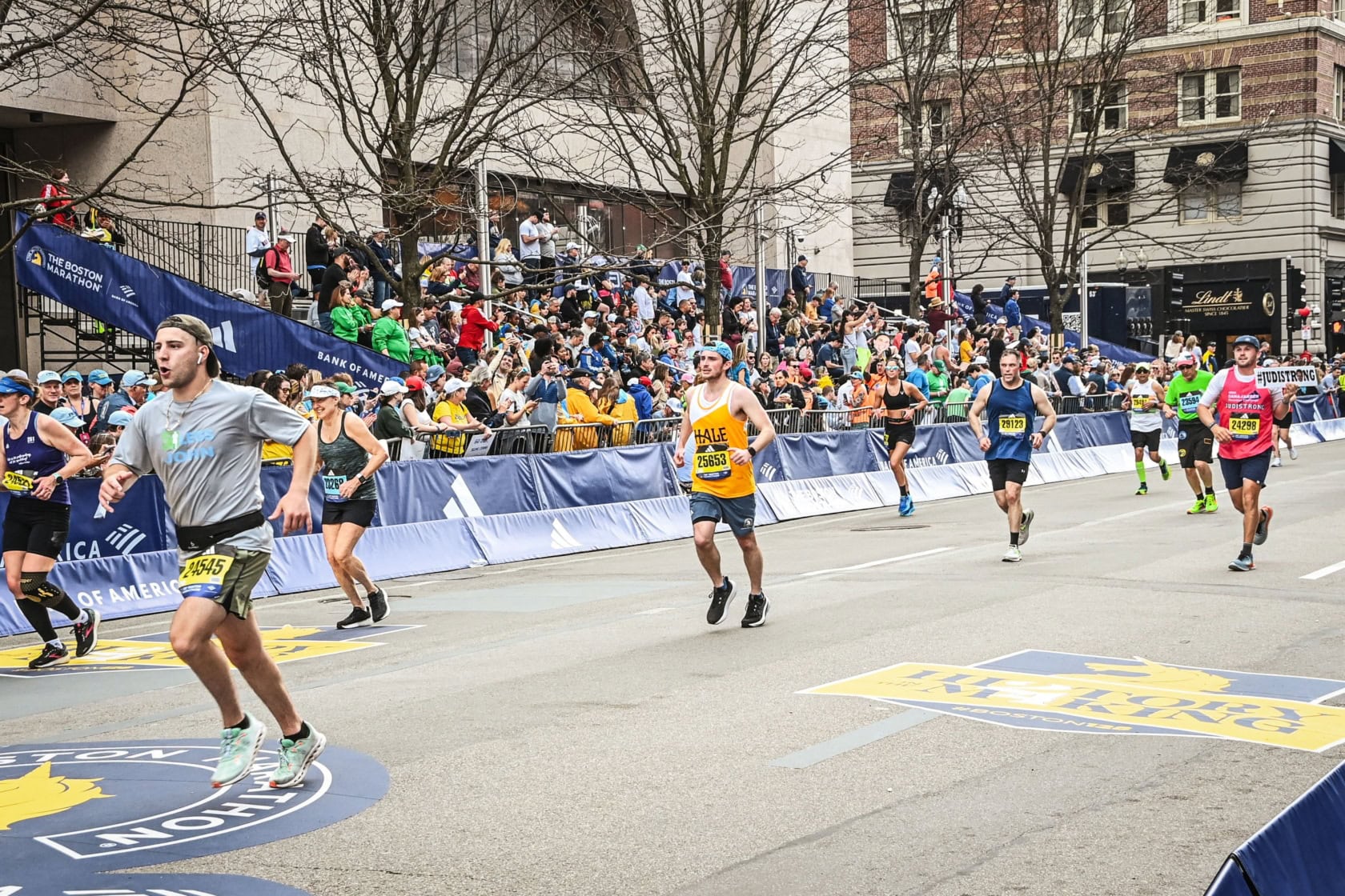 Runners participate in a road race while spectators watch from behind barriers on both sides of the street in an urban setting.