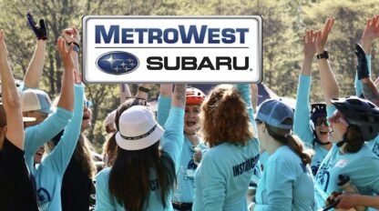 A group of people in blue shirts cheer outdoors under a large MetroWest Subaru logo.