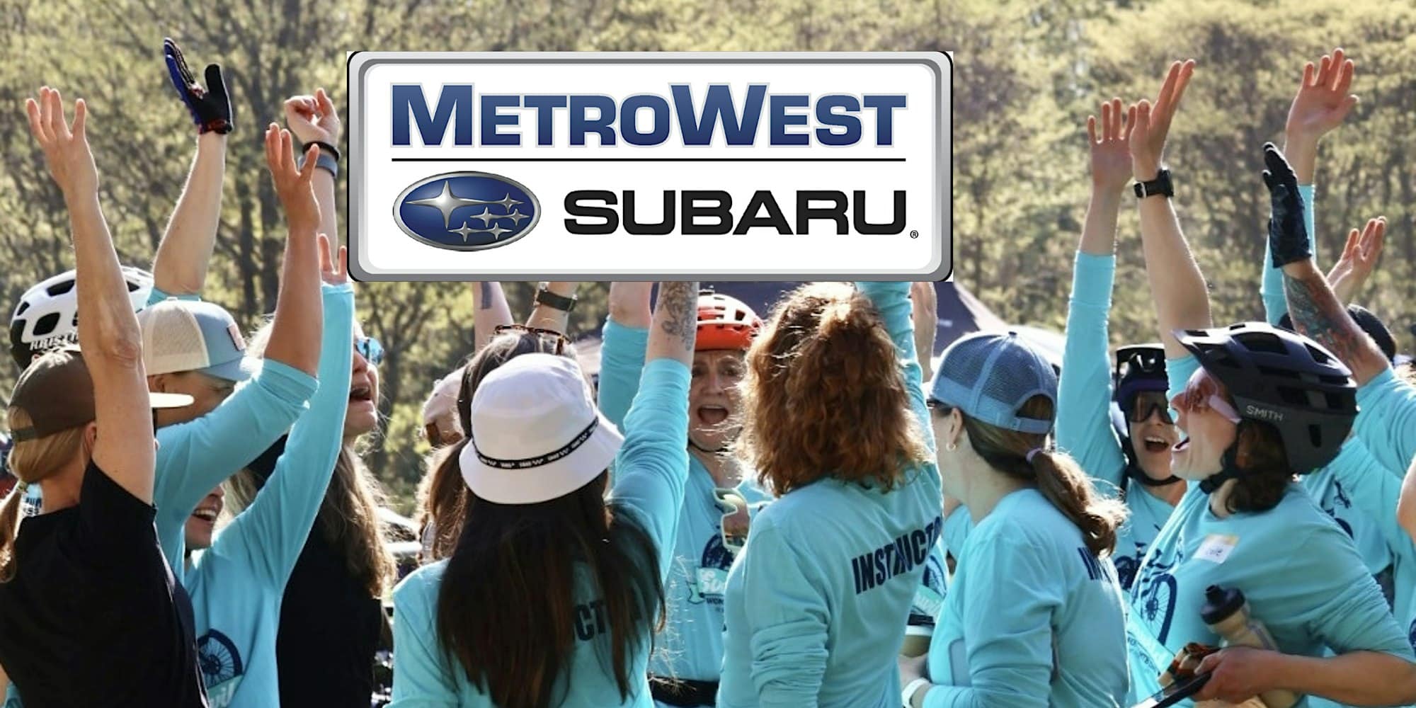 A group of people in blue shirts cheer outdoors under a large MetroWest Subaru logo.